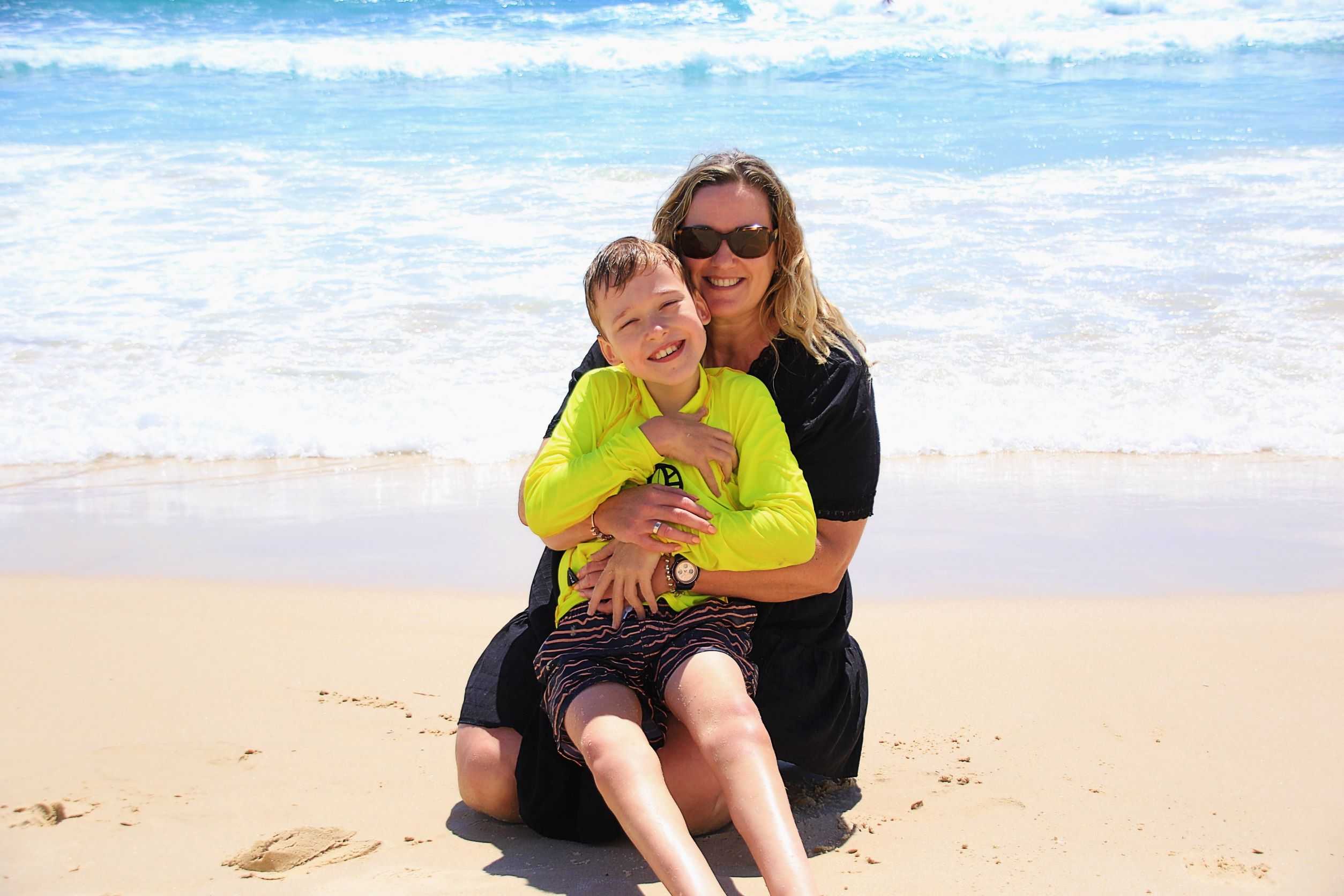 Smiling boy in surfing gear on the lap of his mother who is sitting on the sand by the beach