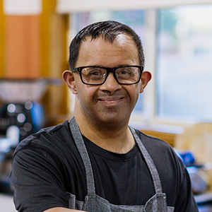NDIS participant, Kaarthik, wearing black glasses and black t-shirt, is smiling at the camera.