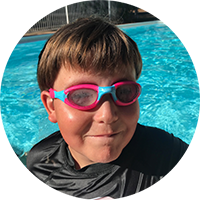 NDIS participant, Christopher, in a pool, wearing pink goggles. Christopher is smiling at the camera. 