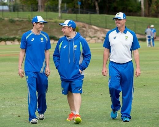 Three men wearing blue cricket uniforms walk across a cricket oval toward the camera.