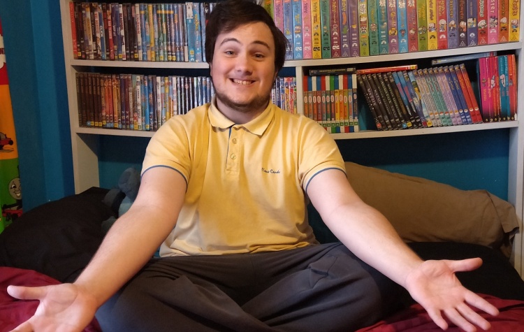 Zac sits cross-legged in front of shelves sagging under the weight of DVDs and videos. He has a huge smile and open arms.