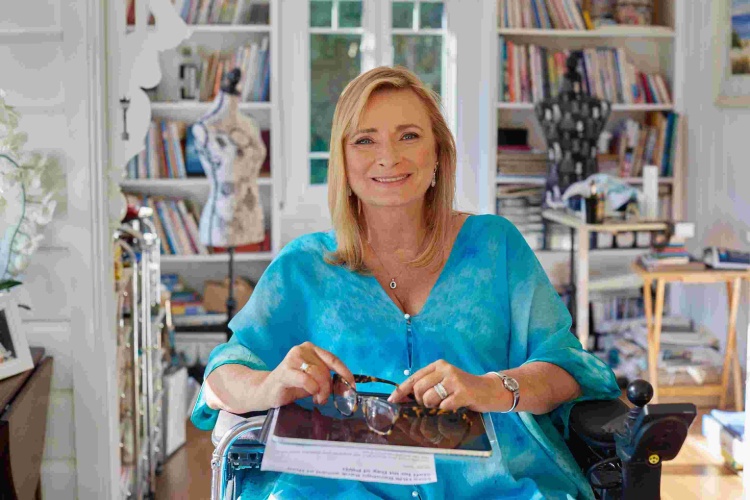 Carol Taylor at home, dressed in a light blue shirt, surrounded by bookshelves and tailor's dummies with designs on them
