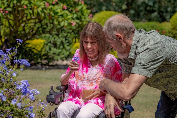 Woman in wheelchair smiles at flower in her hand and her husband leans over her wheelchair arm