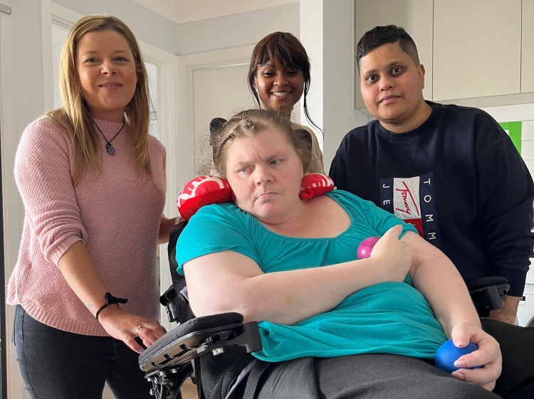 Kelly sitting in wheelchair with two smiling female support workers and her smiling son next to her.