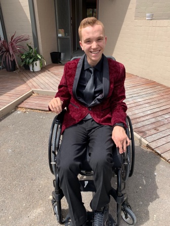 Elijah sits in his wheelchair in a red jacket, ready for his Year 12 Formal