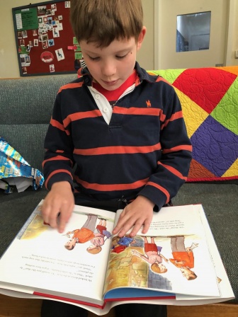 A young boy wearing an orange and black striped jumper sitting on a grey couch next to a colourful quilted blanket reading a picture book.