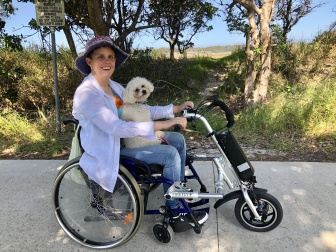 Shelley sitting on her scooter looking at the camera with a small white dog on her lap.