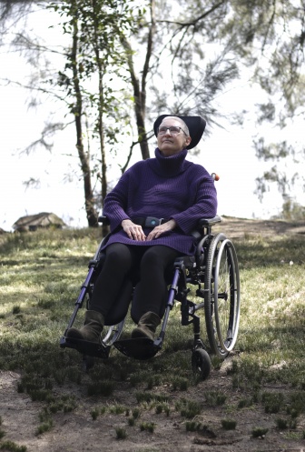 Desney sitting in her wheelchair wearing a purple turtleneck knitted jumper. She is outdoors with trees surrounding her, she is looking up towards them.