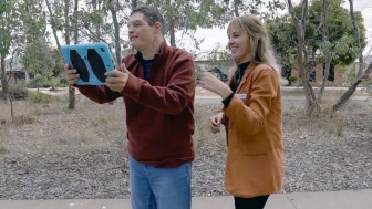 A man is holding up an ipad and looking at the screen. A woman is looking over his shoulder and smiling.