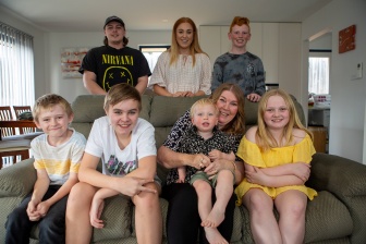 Three teenagers are standing behind a couch. On the couch there are 3 children and a toddler who is sitting on an adult's lap. They're all smiling at the camera. 