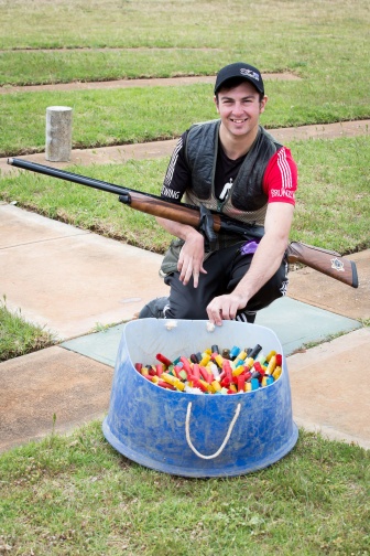 A man is crouching next to a tub of clay shooting markers. He is holding a sports rifle over his knee.