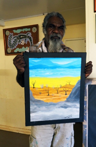 An Aboriginal man is holding a painting of the Australian outback with trees in the foreground and hills in the background.