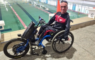 A man in a three wheeled wheelchair is smiling at the camera with an indoor pool in the background.