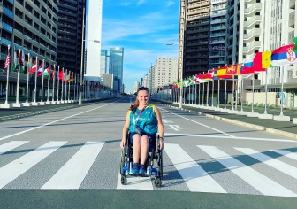 A woman wearing an Australia Olympics top in a wheelchair is in the middle of an empty road. On each side of the road are flags from around the world.