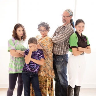 Two adults standing with their arms crossed in an amusing way, they're surrounded by 3 young teenage girls who also have their arms crossed.