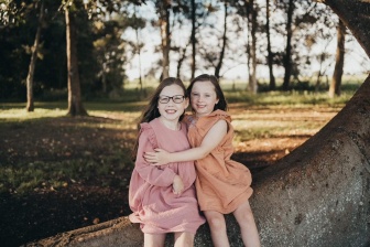 Two young girls are sitting on a tree branch hugging.