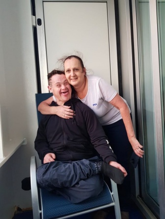 A woman is standing behind a man with Down syndrome sitting in a chair. She has one arm around him in a hug.