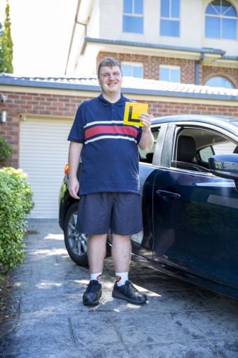 A young man stands in front of a car holding an L plate