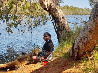 Gaylene sitting on the bank of a river under a tree looking at the camera.