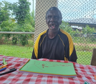 Warren is sitting at a table smiling with a picture that he has drawn in front of him.