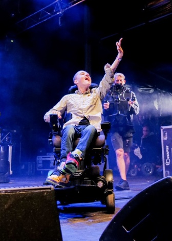 Cooper onstage at Ability fest 2022, raising his arm before the audience. Smoke machines and videographer in the background.