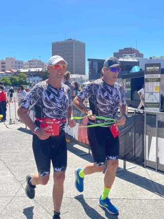 John Domandl and Jason Clark finishing the Ironman race. They are running in tandem against an urban backdrop featuring a blue sky.