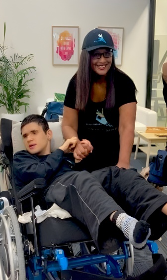 Jaden in his custom wheelchair. A smiling Tanya stands beside him, holding Jaden's hand.