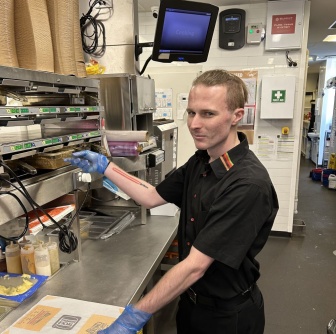 Caleb working serving food at a fast food outlet wearing a black uniform and gloves.