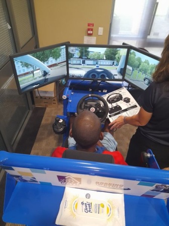 Waylon is sitting in a driving simulator with a steering wheel and three screens.