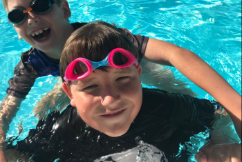 Young male NDIS participant in a pool, wearing pink goggles. Another young male is behind him in the pool with his arms around him. Both are smiling at the camera.