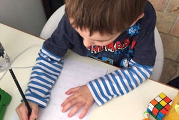 Looking over the top of a six-year-old male NDIS participant and ECEI recipient, practising to write his name at a desk.
