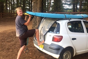 Lleyton unloads his stand up paddle board from the roof of his car. He is barefoot, standing on pine needles and underneath towering Norfolk Island pine trees. He is smiling at the camera.