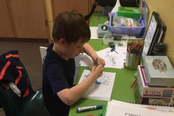 George sitting at a green desk focused on colouring in a picture of Thomas the Tank Engine 