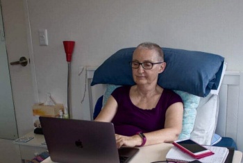 Desney with short hair and glasses sitting up in bed working on a laptop which is sitting on a table over the bed.