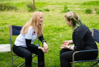 A woman with long blonde hair and wearing a white t shirt is sitting on a blue fold out chair. She is facing the right, leaning forward taking to a woman with green hair highlights. 