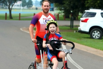 A young girl in a sports suit is riding a large tricycle. A man in a matching suit runs behind holding onto the handles.