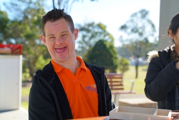 A man with Down syndrome is sitting at an outdoor table and smiling at the camera.