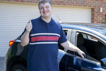 A young man stands in front of a car giving a thumbs up.
