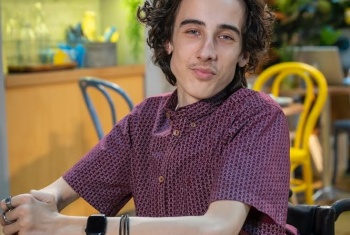 Oli sits at a table, with his arms resting on it, looking relaxed, on the Neighbours set