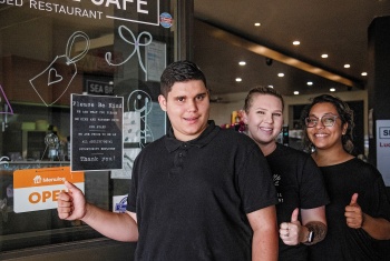 A young man cafe standing in front of cafe entrance with two smiling women next to him. All three have their thumbs up.