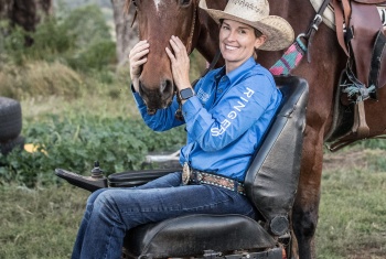 Smiling woman wearing a riding hat and sitting in wheelchair stroking her horse