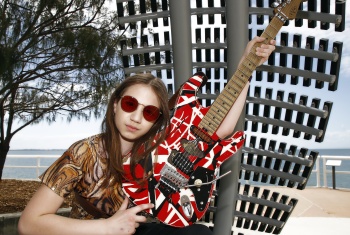 Young woman with long brown hair and sunglasses holding her guitar. In the background the beach can be seen