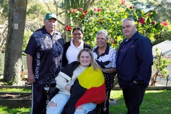 A smiling Tanesha siting with an Aboriginal flag draped over her shoulders. Her family stand around her, against a backdrop of  flowering trees.