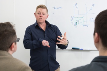 Paul standing in front of a whiteboard with a graph on it - Relationships on the horizontal axis and systems on the vertical axis. Two audience members are oaying close attention to him. 
