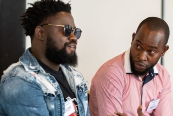 Sengo wearing a denim jacket and sunglasses sitting next to a man in a red shirt who is talking to him. They're both wearing event style name badges.