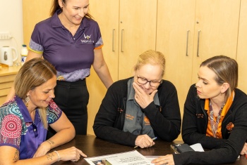 3 women sit around a table looking at some papers, one stands with them.